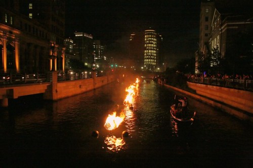 A river at night with flames floating on the water, and a gondola passing by under city lights.