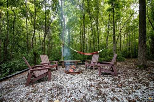 A cozy campsite with four wooden chairs around a fire pit, surrounded by trees and a hammock hanging nearby.