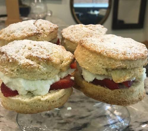 Four strawberry shortcakes with whipped cream, dusted with powdered sugar, displayed on a glass stand.