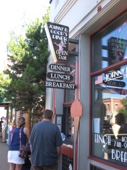Sign for Johnny B. Good's Diner displaying hours and meal options: breakfast, lunch, and dinner. People in line outside.