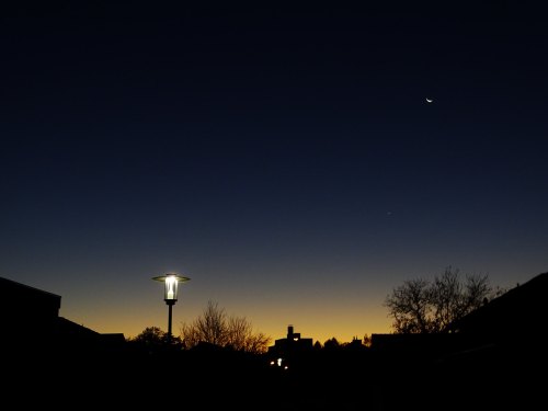 Silhouette of buildings and trees against a twilight sky, with a crescent moon and a bright star visible.