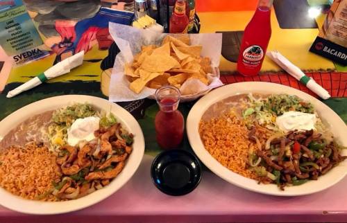 Two plates of Mexican food with rice, beans, grilled meat, and toppings, accompanied by a bowl of tortilla chips and salsa.