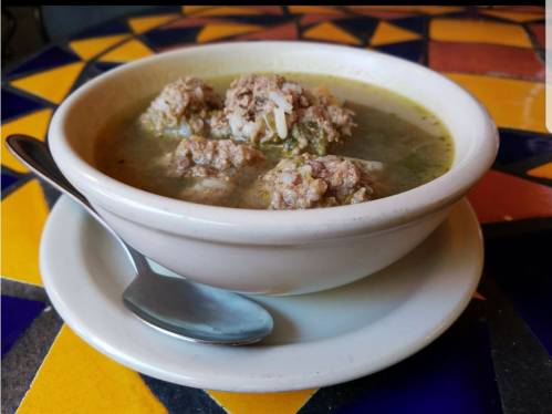 A bowl of soup with meatballs, served on a colorful tiled table, with a spoon beside it.