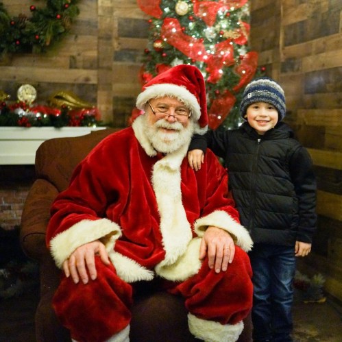 A smiling child stands next to Santa Claus in a cozy, festive setting with a decorated Christmas tree.