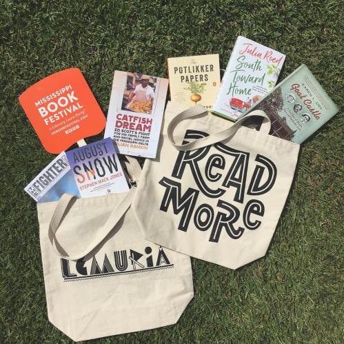 Two canvas tote bags on grass, one with "READ MORE" printed, surrounded by books and a flyer for a book festival.