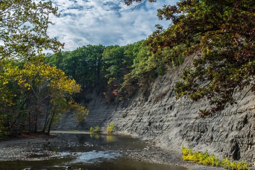 A serene river flows beside a steep, rocky cliff, surrounded by lush trees and a partly cloudy sky.