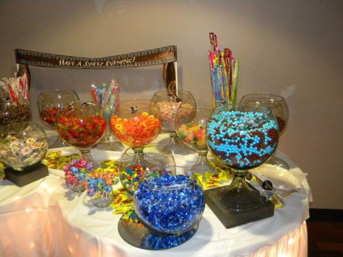 A colorful candy buffet featuring various candies in glass bowls, with a sign that says "Have a Sweet Evening."