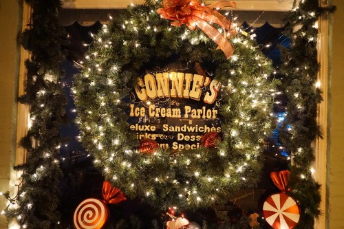 A festive wreath with lights and candy decorations surrounds a sign for Connie's Ice Cream Parlor.