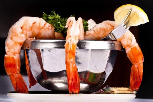 A silver bowl filled with shrimp, garnished with parsley and a lemon wedge, resting on a white plate.