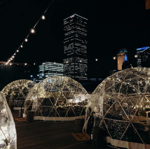 A nighttime view of illuminated igloo-like structures with city skyscrapers in the background, adorned with string lights.