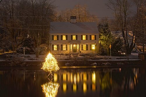 A snowy house at night, illuminated by warm lights, with a glowing Christmas tree reflected in the water.