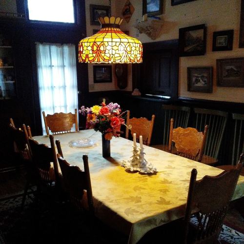 A cozy dining room with a large table, floral centerpiece, and a stained glass lamp hanging above.
