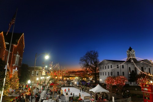 A festive town square at dusk, adorned with holiday lights and decorations, featuring an ice skating rink and a clock tower.