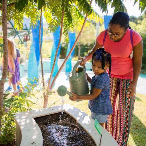 A woman and a child water plants in a garden, surrounded by greenery and colorful decorations.