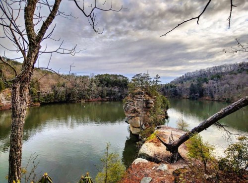 A serene lake surrounded by trees and rocky cliffs under a cloudy sky, reflecting the landscape in the water.