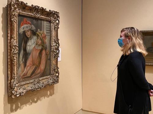 A visitor in a mask observes a painting of two girls in a museum, framed in an ornate gold frame.