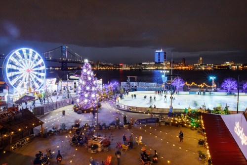 A festive waterfront scene with a skating rink, a lit Christmas tree, a Ferris wheel, and city skyline at night.