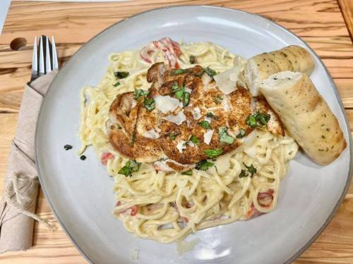 A plate of creamy pasta topped with seasoned chicken, herbs, and a side of garlic bread on a wooden table.