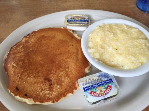 A plate with a golden pancake, a bowl of grits, and two butter packets on the side.