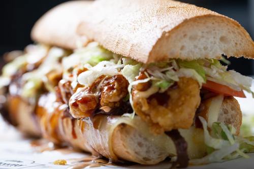 A close-up of a loaded sandwich with crispy chicken, lettuce, tomato, and drizzled sauce in a toasted hoagie roll.