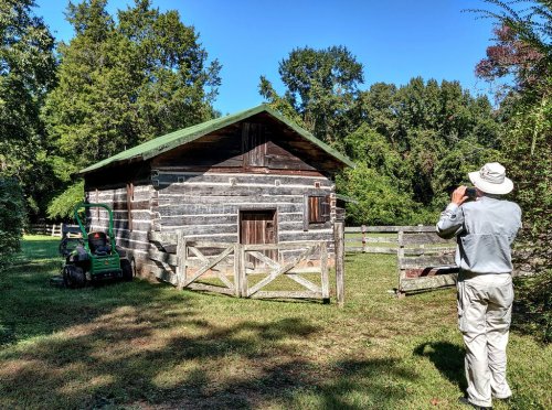 A person in a hat takes a photo of a rustic log cabin surrounded by trees and a lawn mower nearby.