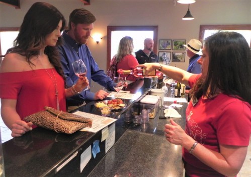 A woman in a red top pours wine for a customer at a bar, while another woman and a man look on. Snacks are on the counter.