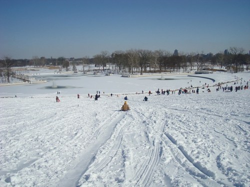 A snowy hill with people sledding and playing, surrounded by trees and a frozen pond under a clear blue sky.