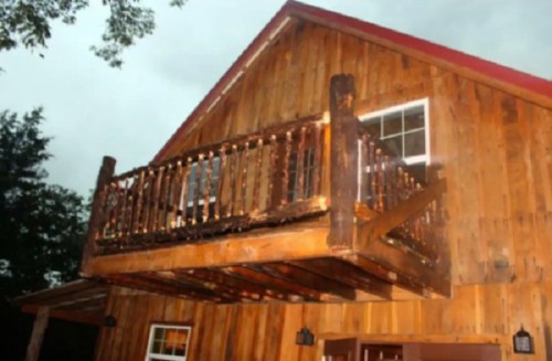 A wooden cabin with a balcony, featuring a rustic design and a red roof, set against a cloudy sky.