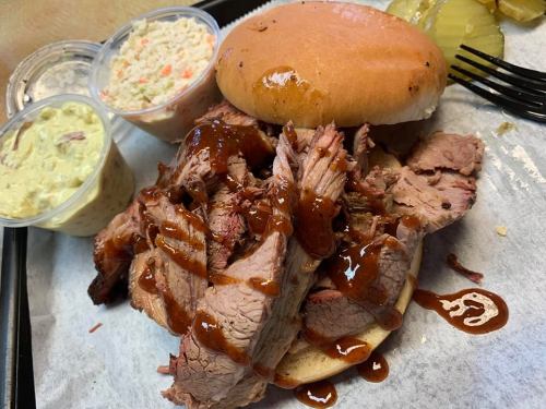 A close-up of a BBQ brisket sandwich topped with sauce, served with coleslaw and pickles on the side.