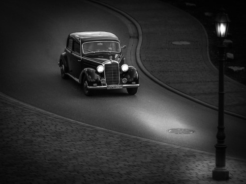 A vintage black car drives along a curved, cobblestone road, illuminated by a streetlamp in black and white.