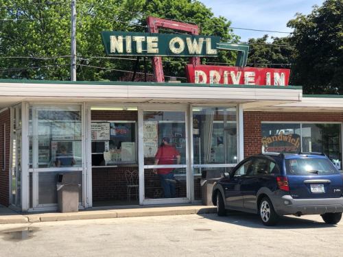 A retro drive-in restaurant with a neon sign reading "Nite Owl Drive In" and a parked car in front.