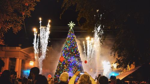 A festive Christmas tree adorned with lights and a star, surrounded by fireworks and a cheering crowd at night.