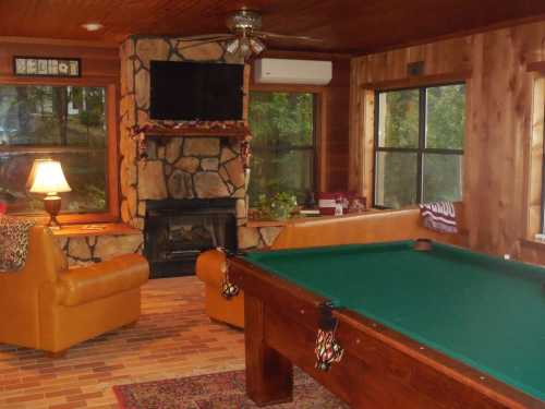 Cozy room with a pool table, stone fireplace, TV, and comfortable seating by large windows. Warm, inviting atmosphere.