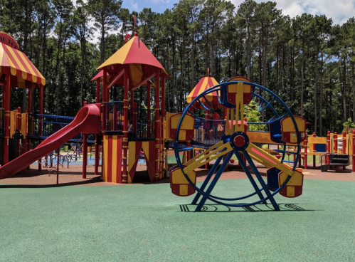 Colorful playground with a slide, climbing structures, and a small Ferris wheel, surrounded by trees and blue sky.