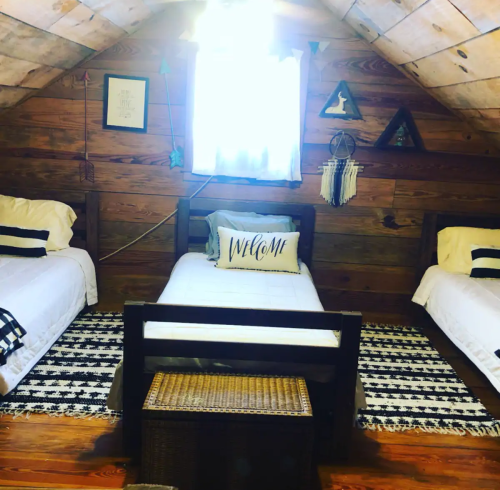 Cozy attic bedroom with three beds, wooden walls, a welcome pillow, and a bright window.
