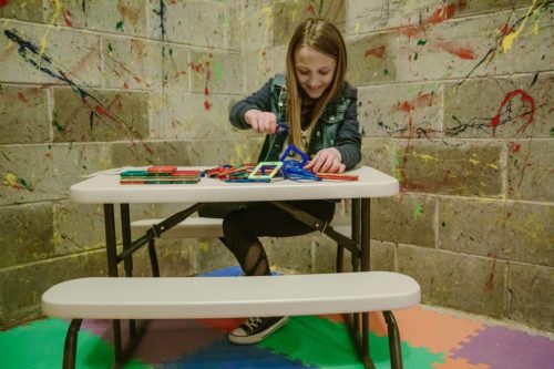 A young girl sits at a table, playing with colorful building blocks in a creatively painted room.