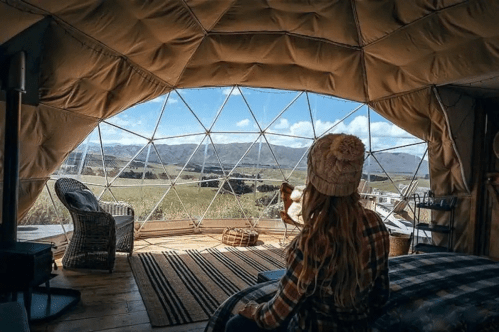 A person sits inside a cozy dome-shaped room, gazing out at a scenic landscape through large, geometric windows.