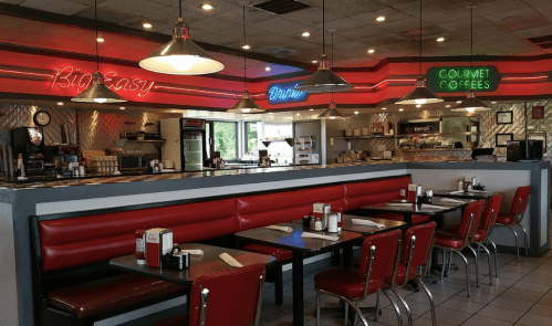 A retro diner interior with red booths, neon signs, and a counter displaying food and drinks.