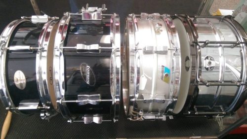 A row of four snare drums in various colors and finishes, showcasing their chrome hardware and distinct designs.
