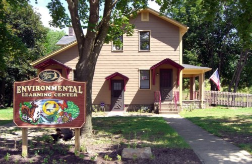 A brown house with a sign reading "Environmental Learning Center" and an American flag, surrounded by trees and grass.