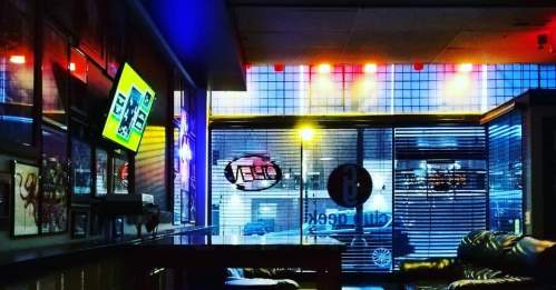 A cozy interior of a bar with colorful lighting, a TV on the wall, and large windows showing a city view.
