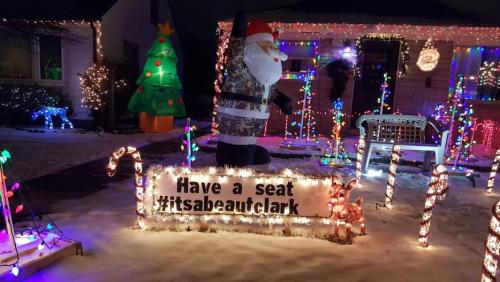 A festive yard display featuring a Santa in camouflage, colorful lights, a Christmas tree, and a sign that says "Have a seat."