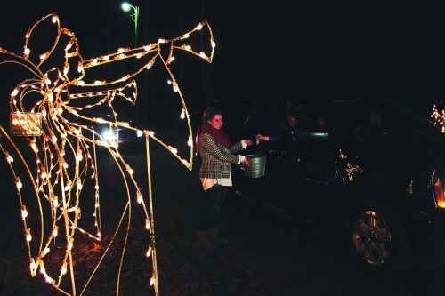 A woman hands a bucket to someone in a car, surrounded by festive holiday lights at night.