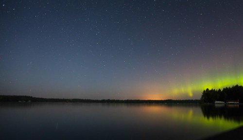 A serene lake at night reflecting a starry sky and vibrant auroras, with silhouettes of trees along the shore.