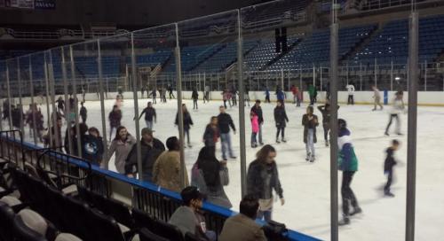 A crowded ice skating rink with many skaters gliding on the ice, surrounded by empty seating.