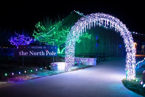 A brightly lit archway welcomes visitors to the North Pole, surrounded by colorful lights and trees at night.