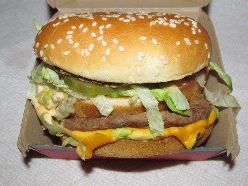 A sesame seed bun sandwich with lettuce, cheese, and a meat patty, served in a cardboard box.