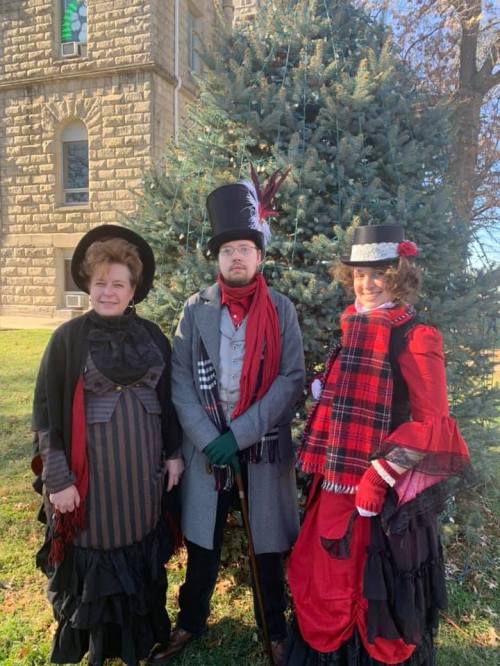 Three people in vintage holiday attire stand in front of a Christmas tree, smiling in a festive outdoor setting.