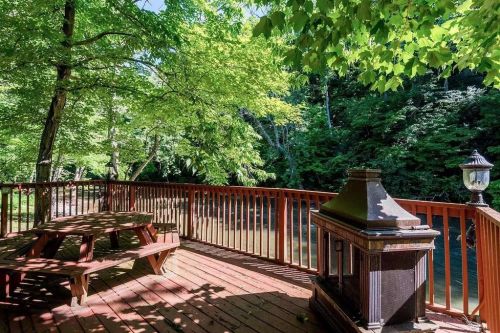 A wooden deck with a picnic table and lantern, surrounded by lush green trees and a calm river.