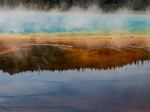 A vibrant geothermal pool with blue water and steam, surrounded by colorful mineral deposits and a forest reflection.